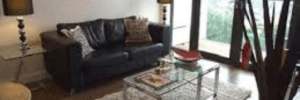 Living room with a James Bond themed picture, black leather sofa, brown flooring, glass table on top of a white rug