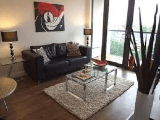 Living room with a James Bond themed picture, black leather sofa, brown flooring, glass table on top of a white rug